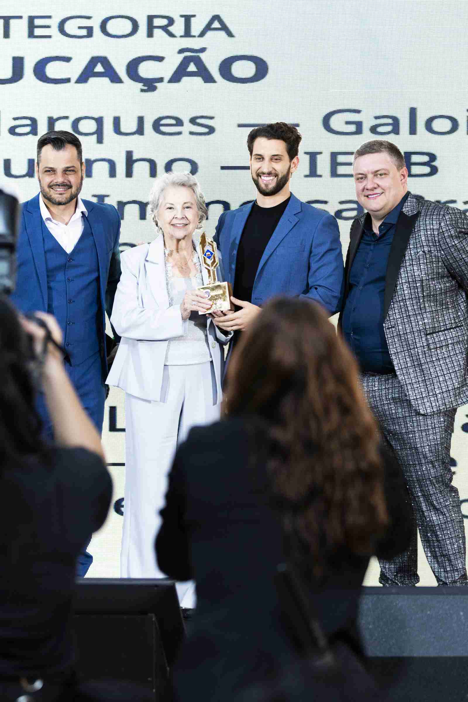 Quatro premiados sorridentes seguram o troféu dourado no palco durante a entrega da categoria Educação, com o telão exibindo os nomes dos indicados ao fundo