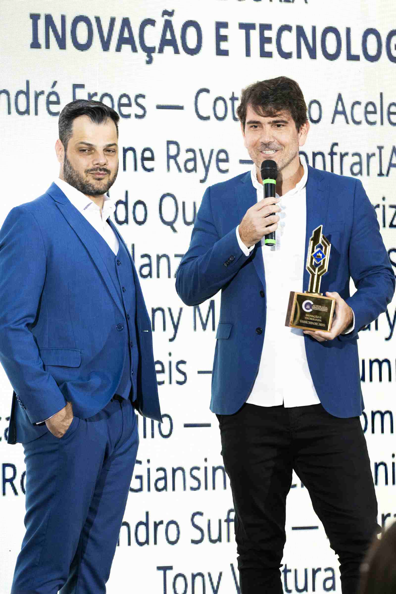 Vencedor da categoria Inovação e Tecnologia discursa ao microfone segurando o troféu, ao lado de outro homem de terno azul, com o telão da categoria ao fundo