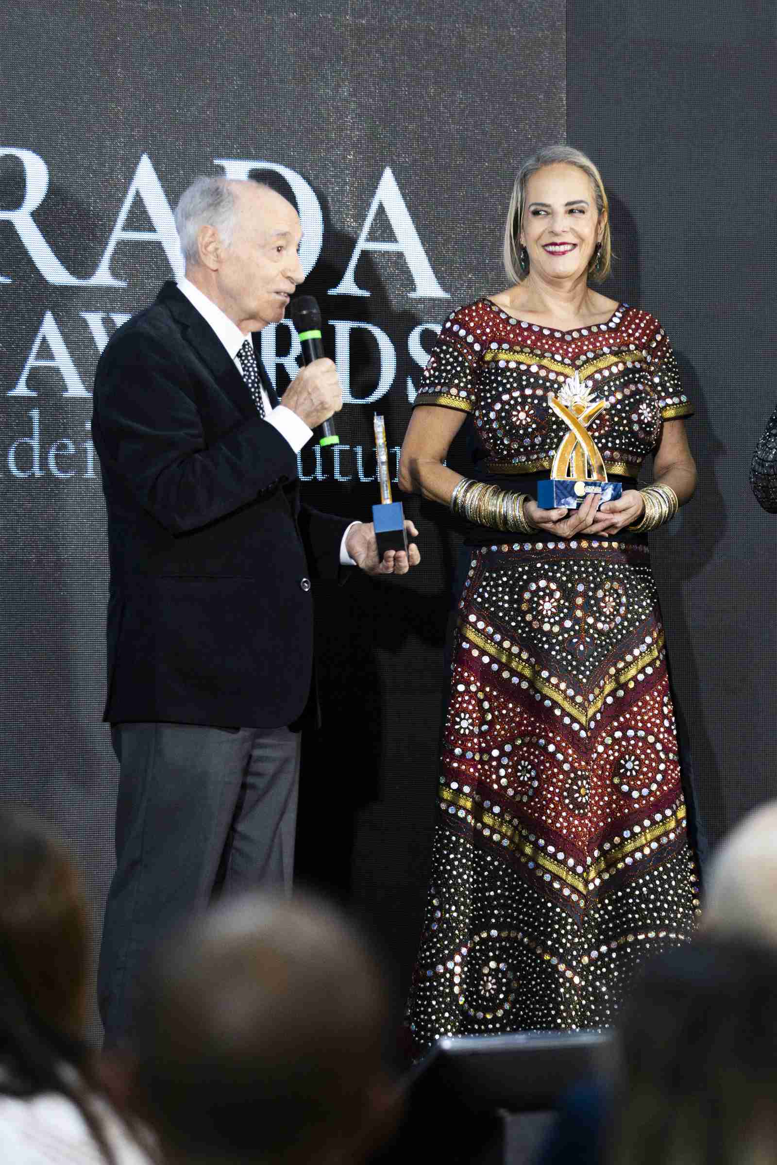 Mulher em vestido de festa bordado dourado e vermelho recebe o troféu das mãos de um apresentador de terno preto, com o letreiro 'ALVORADA AWARDS' luminoso atrás