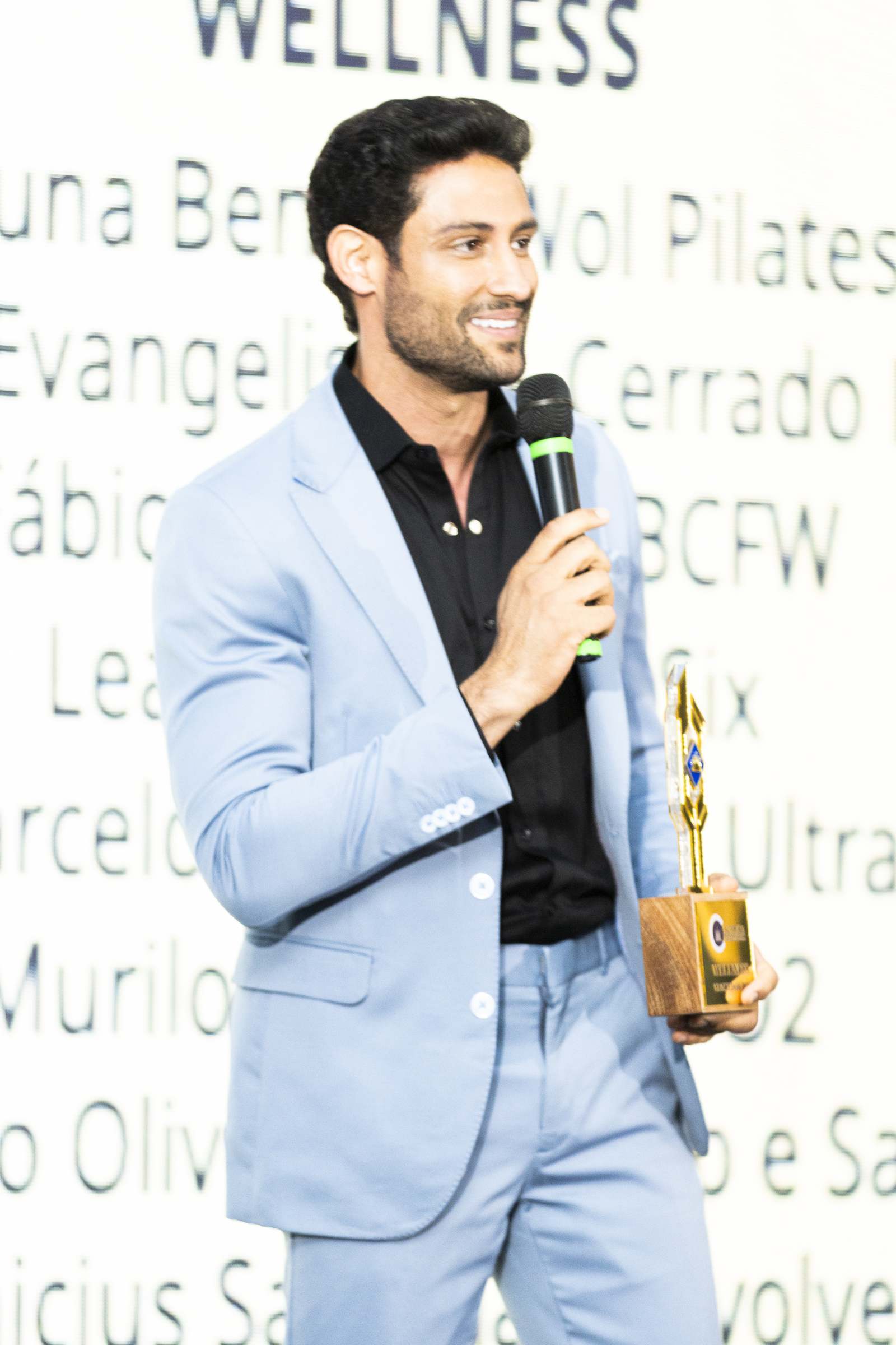 Vencedor da categoria Wellness, de terno azul-claro, sorri segurando o troféu e o microfone diante do telão com a lista de indicados