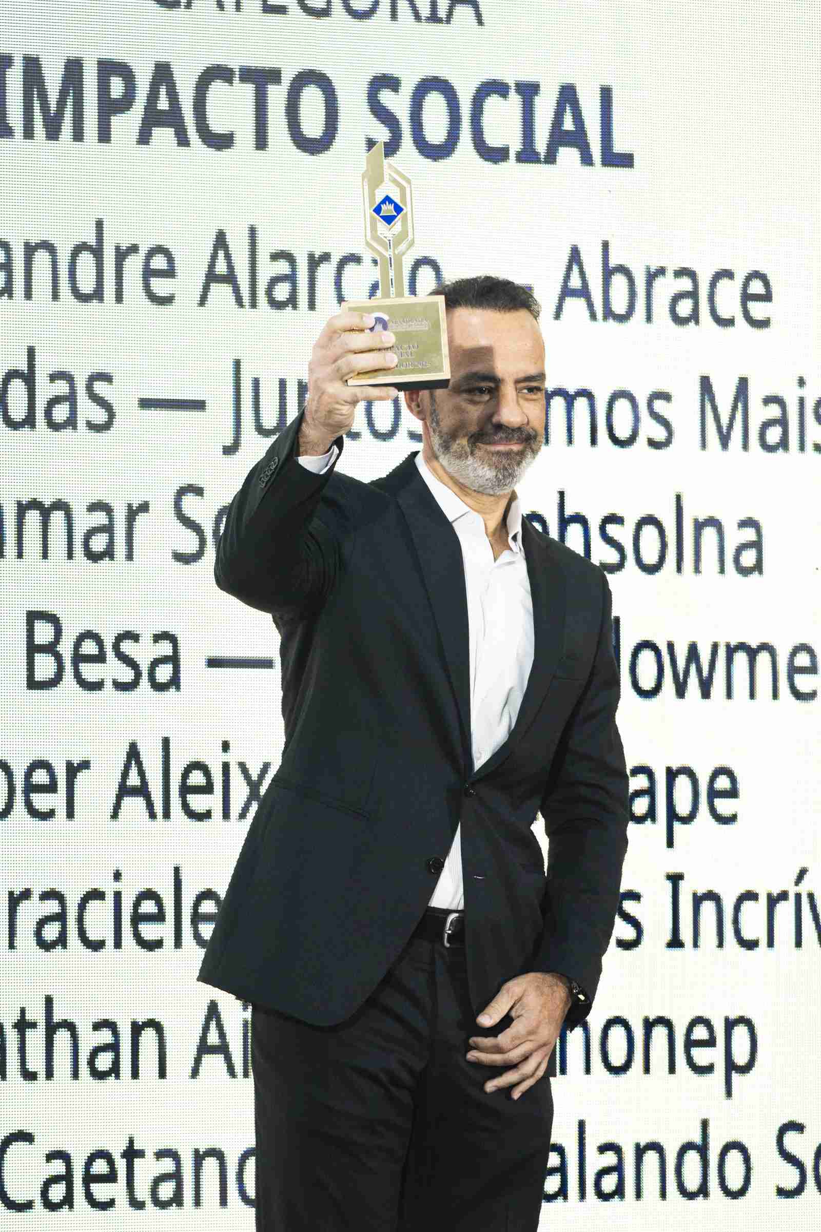 Homem de terno preto ergue o troféu da categoria Impacto Social com o braço estendido, sorrindo, diante do telão com os nomes dos indicados