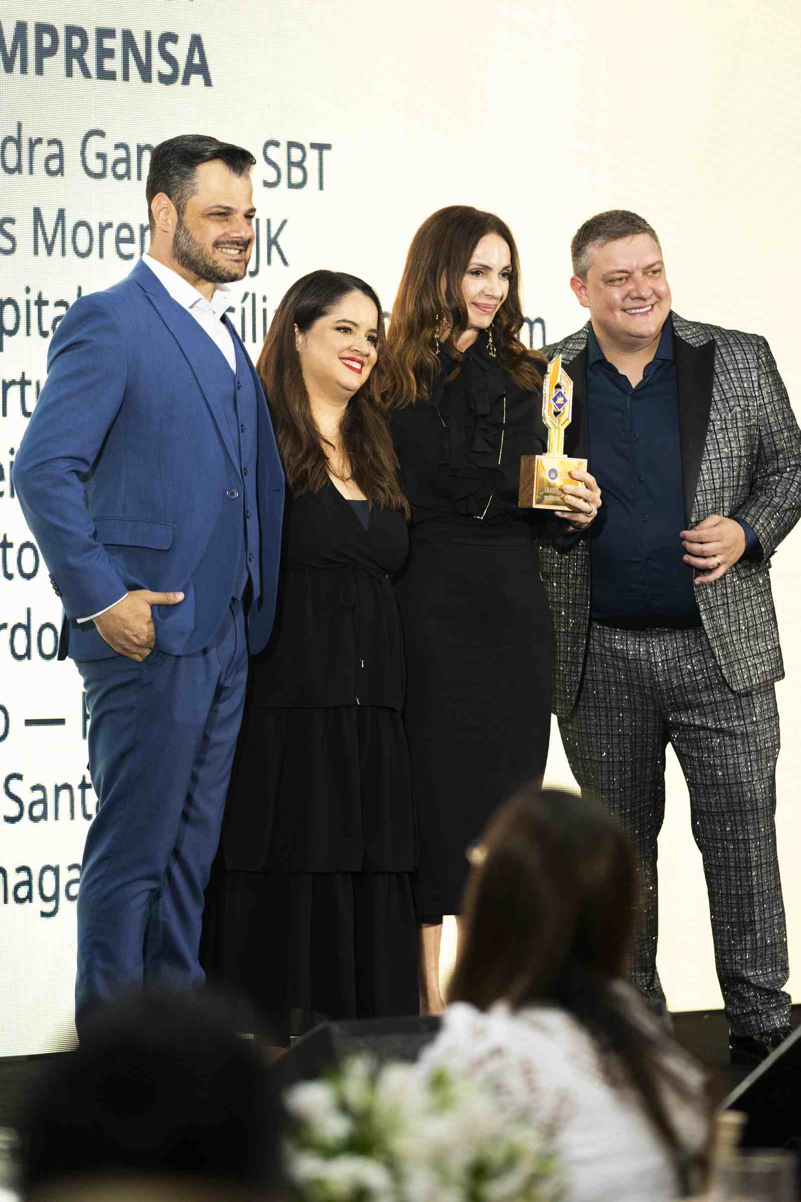 Quatro premiados da categoria Imprensa posam no palco com o troféu dourado em mãos, diante do telão com a lista de indicados