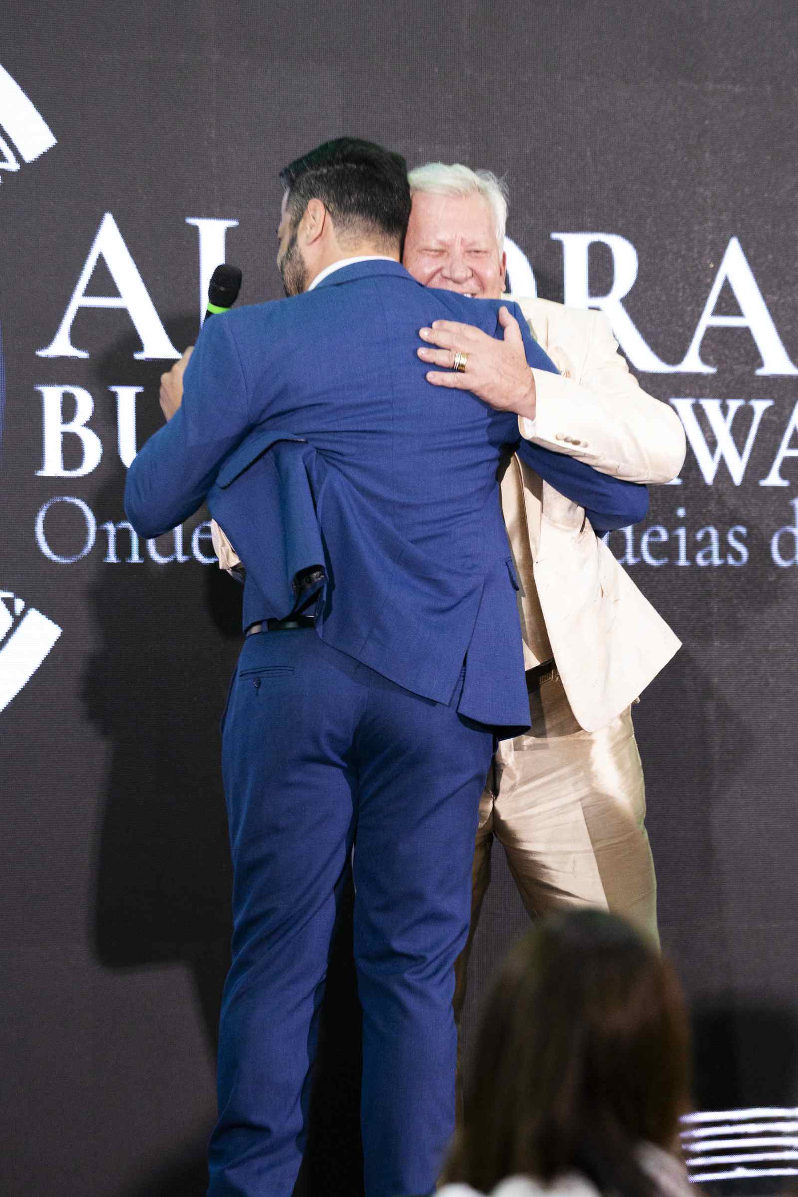 Dois homens se abraçam emocionados no palco, um deles com o microfone na mão, diante do letreiro 'ALVORADA BUSINESS AWARDS'