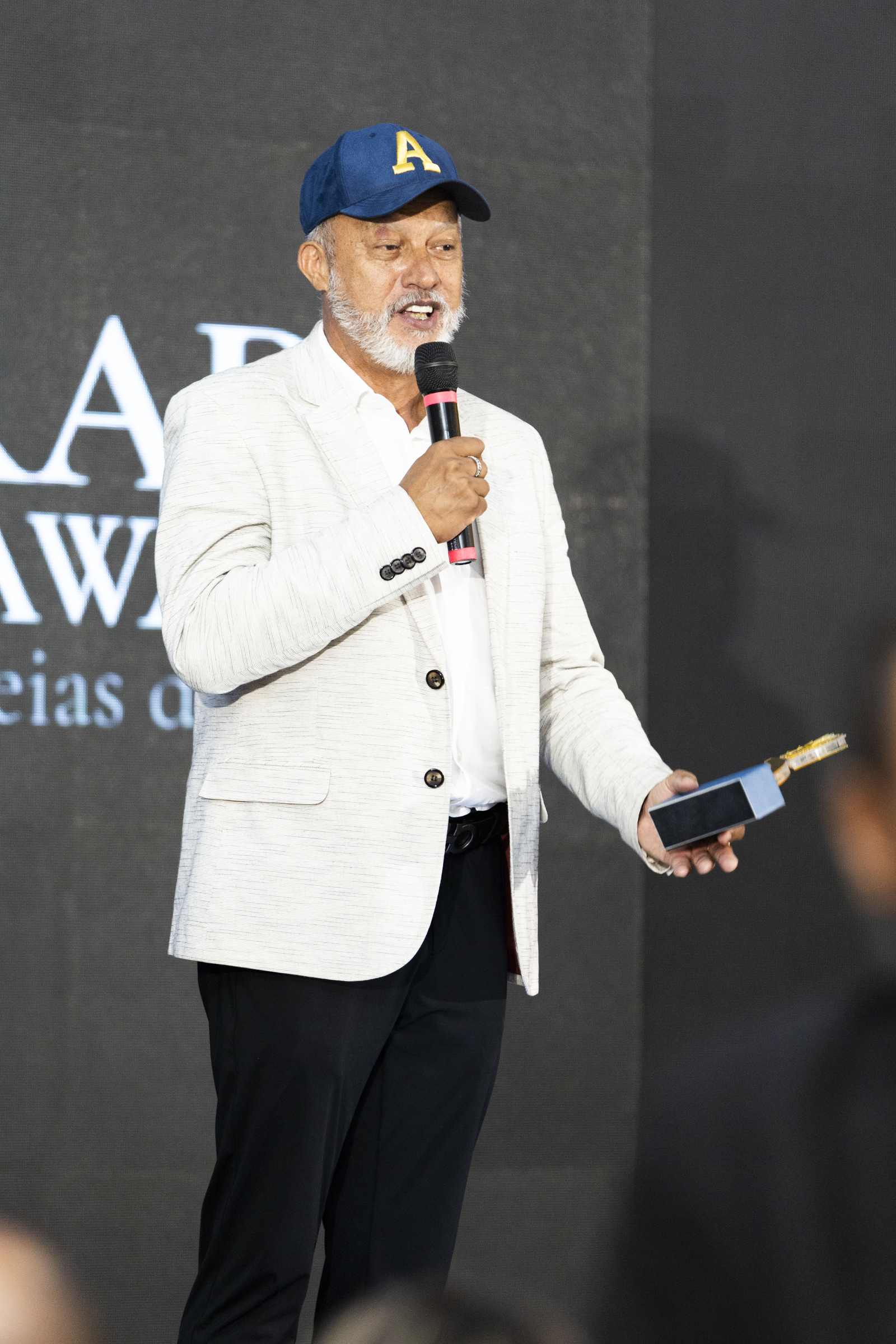 Homem de boné azul e blazer bege discursa sorrindo ao microfone no palco do Olivier Hall