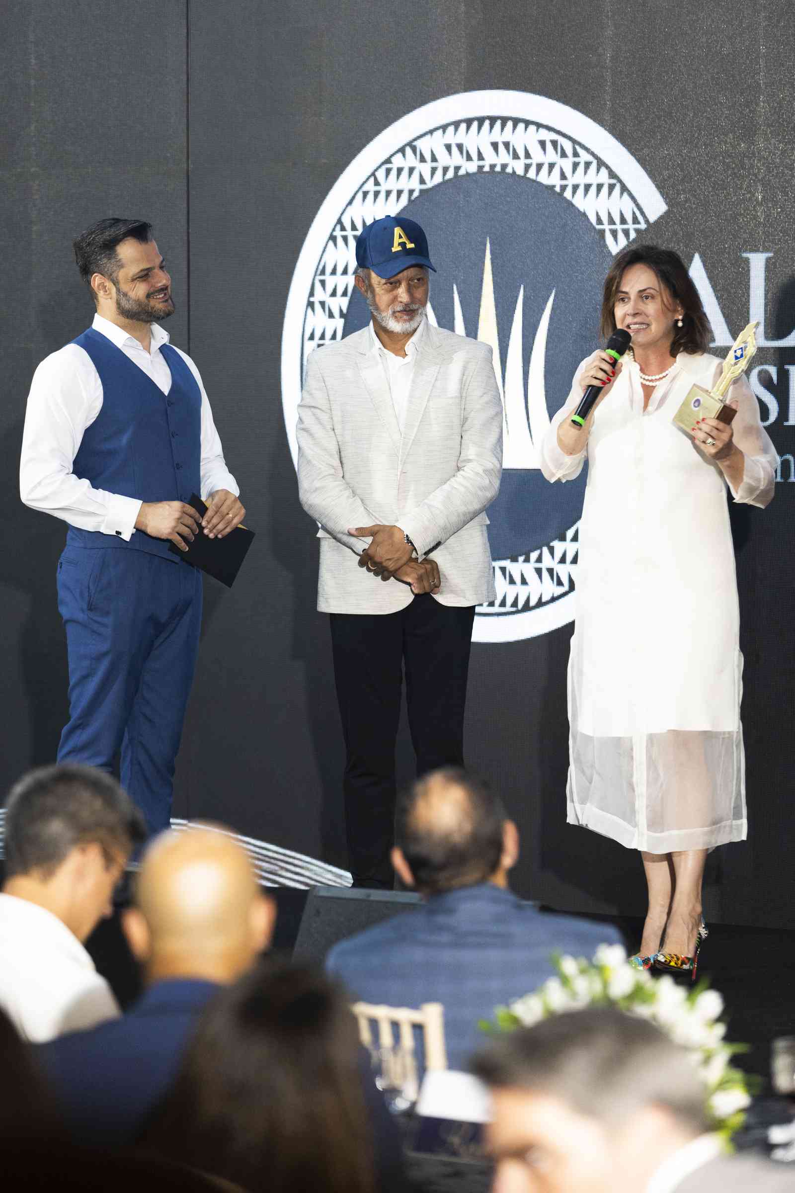 Mulher de vestido branco discursa ao microfone segurando o troféu, ladeada por dois homens no palco, com o símbolo circular do Alvorada Business Awards ao fundo