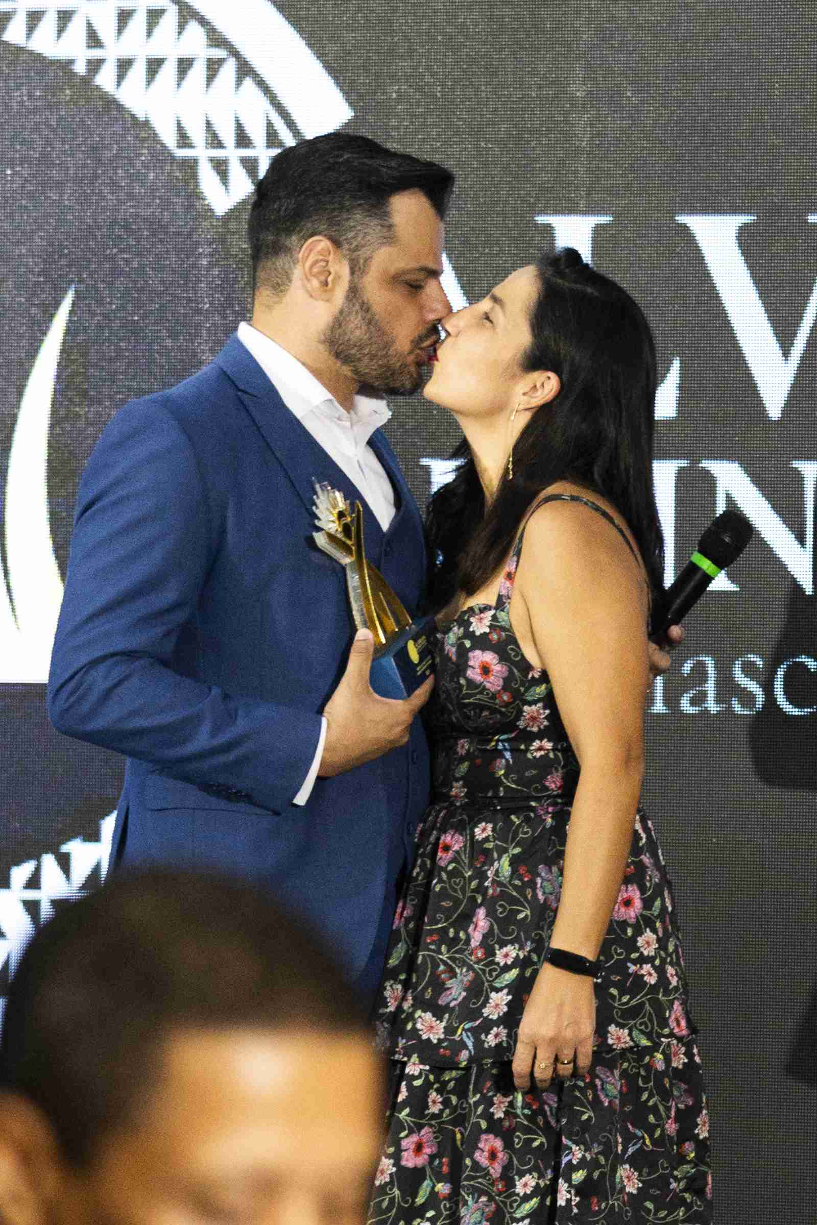 Casal se beija no palco após a entrega do troféu, que o homem segura preso ao paletó, diante do letreiro do Alvorada Business Awards