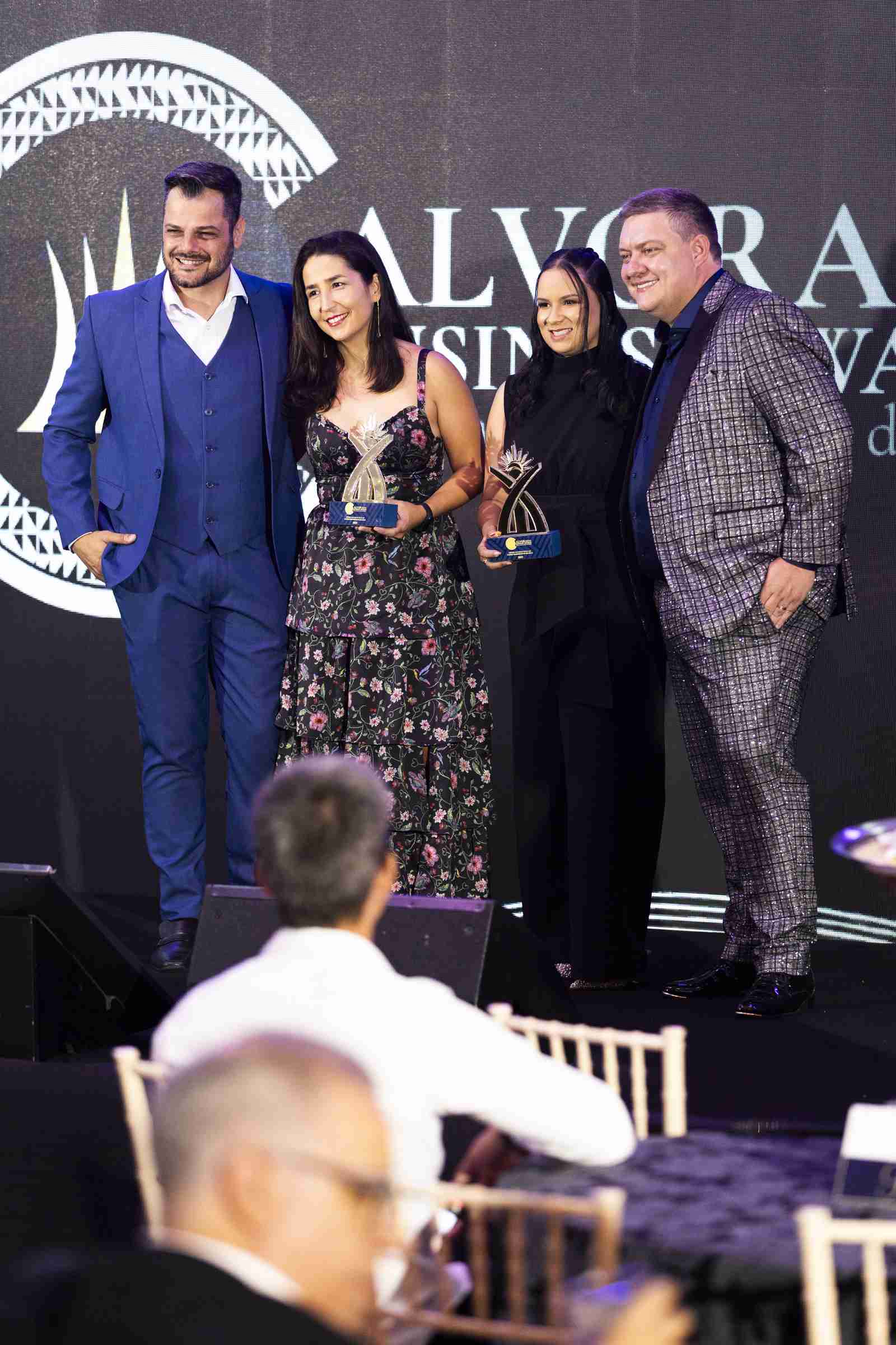 Duas premiadas seguram troféus de cristal no palco, sorrindo ao lado de outras duas pessoas, diante do círculo do logotipo do Alvorada Business Awards