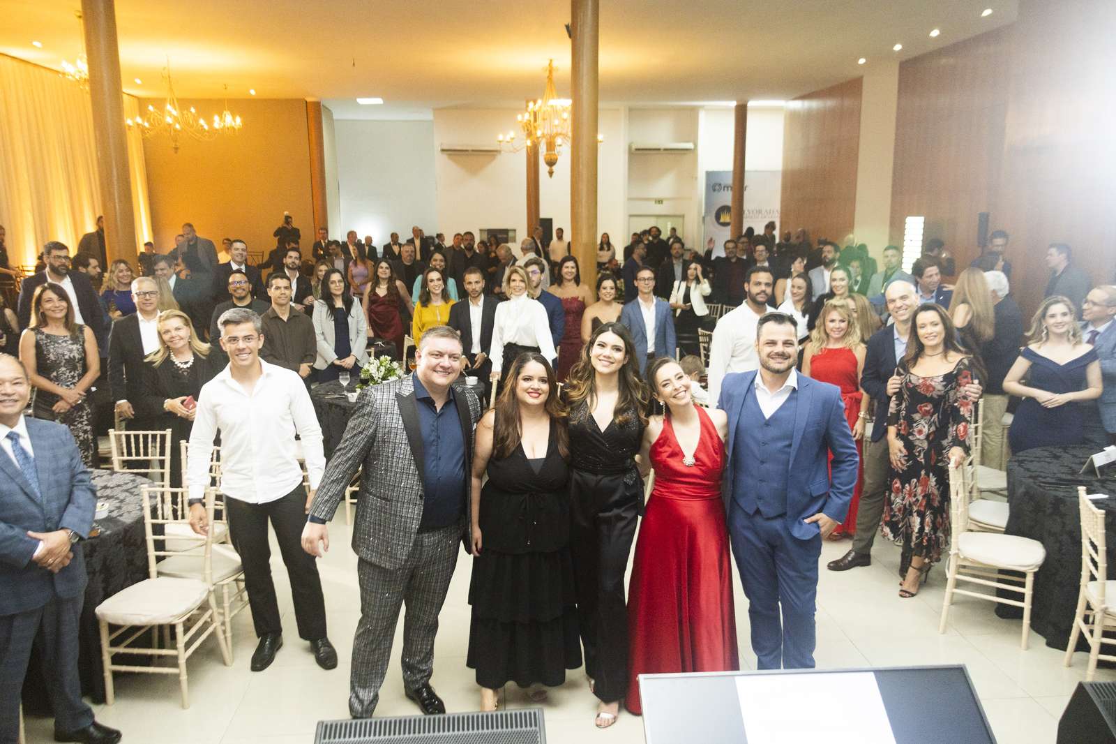 Grupo de convidados posa sorrindo em primeiro plano no salão do Olivier Hall, com lustres dourados e a plateia completa ao fundo