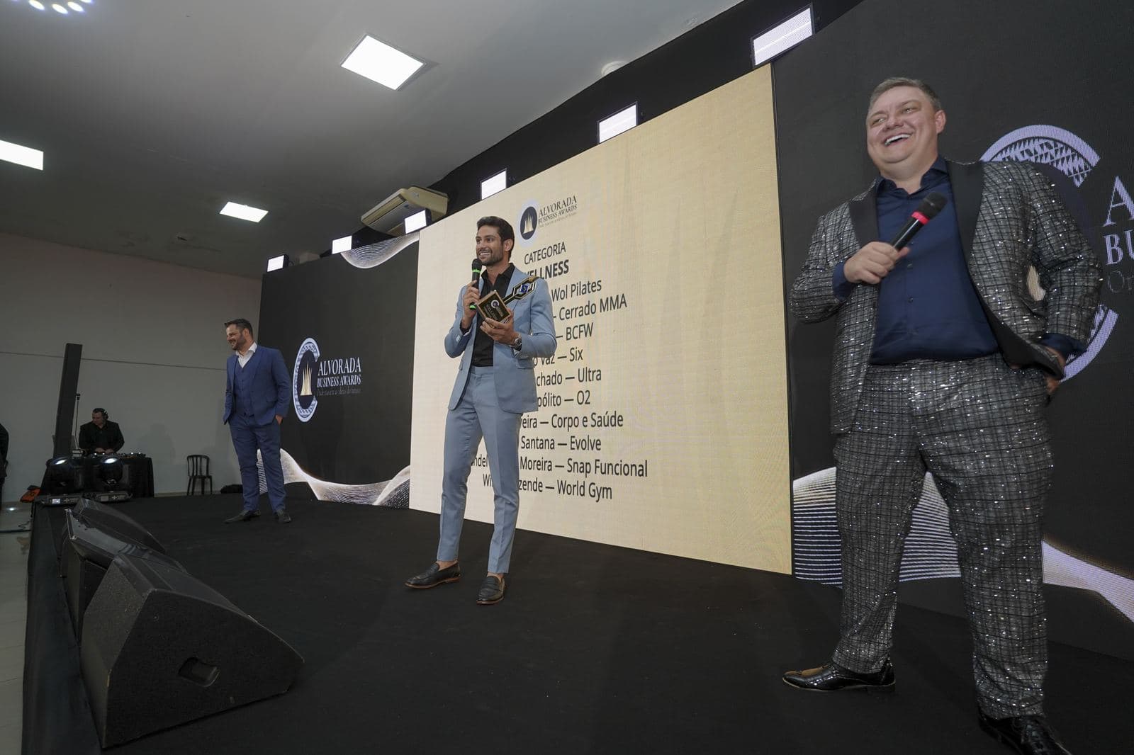 Visão ampla do palco com três apresentadores durante o anúncio da categoria Wellness, mostrando a escala do salão do Olivier Hall