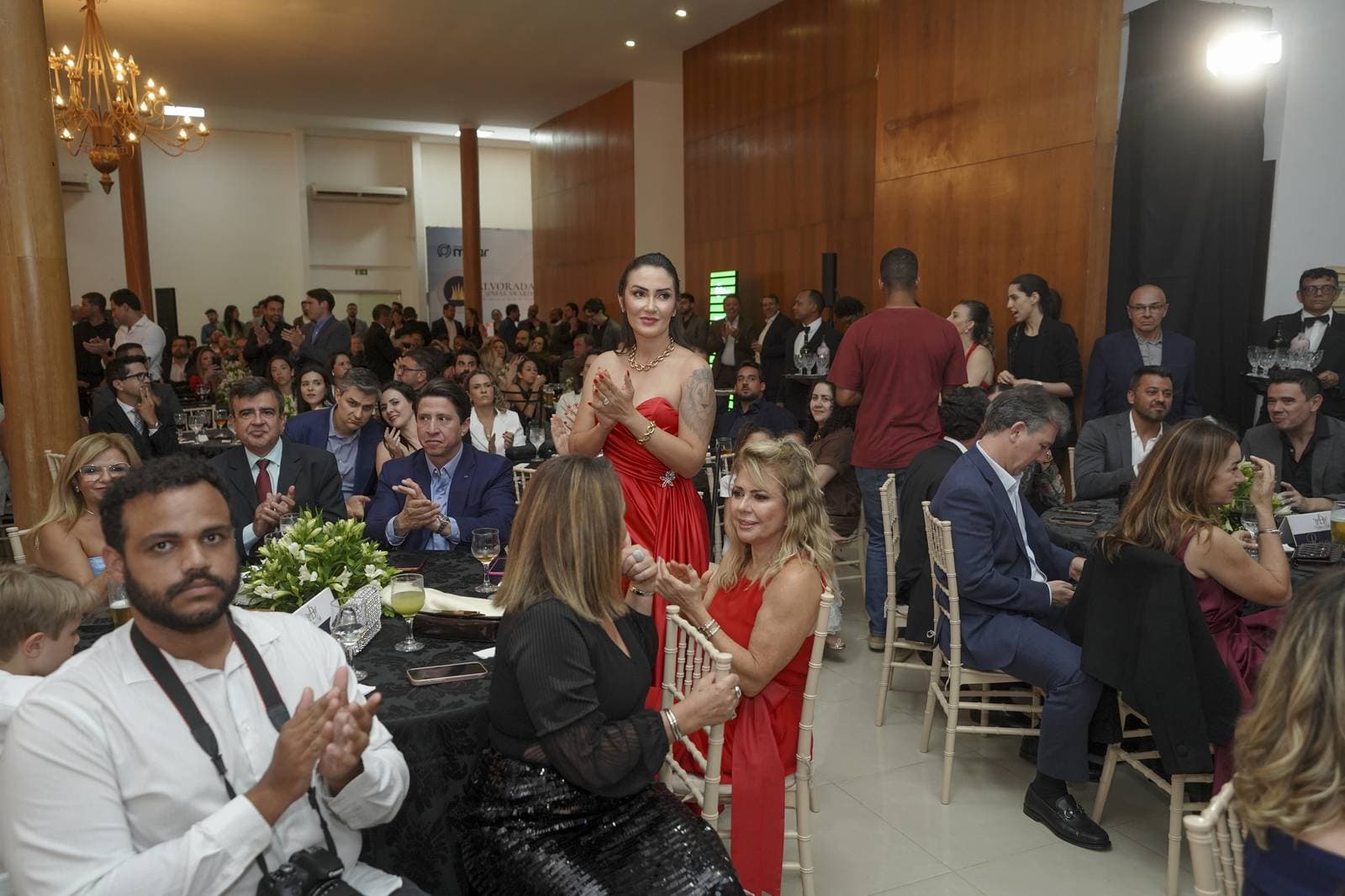 Convidada de vestido vermelho aplaude em pé em meio à plateia lotada do jantar de gala