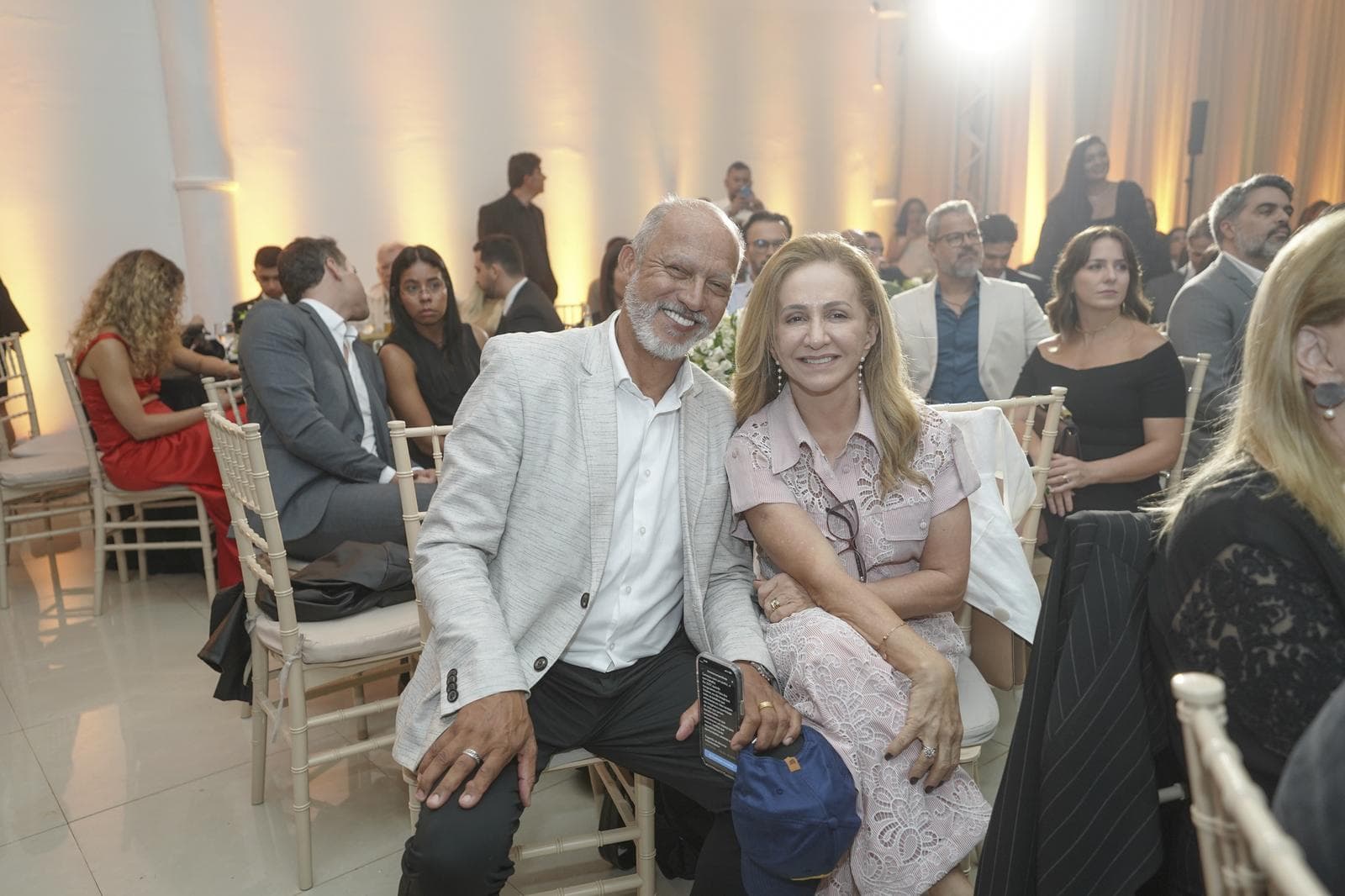 Casal de convidados sorri abraçado em meio à plateia sentada, com o salão do evento iluminado em tons dourados ao fundo