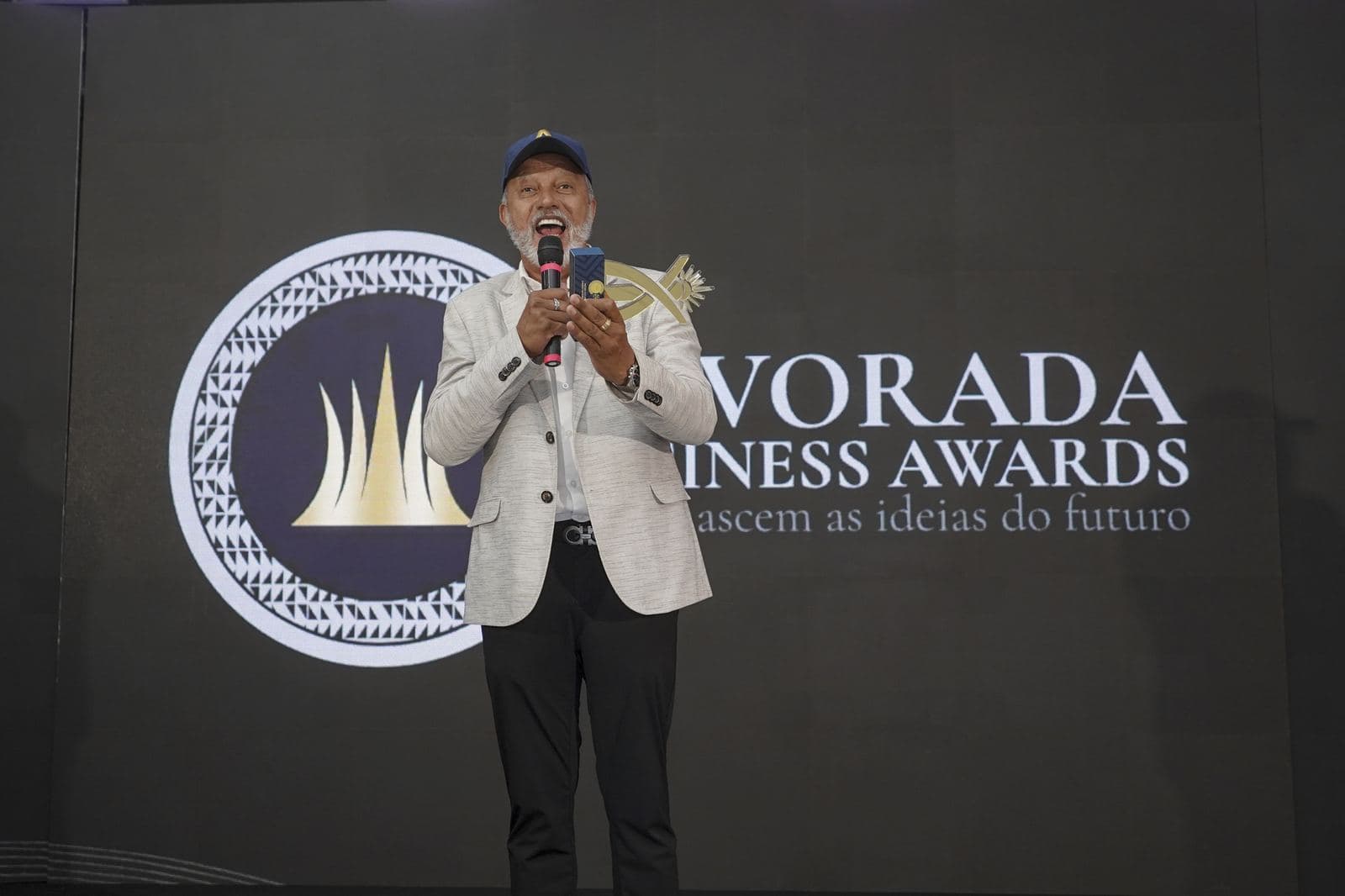 Homem de boné discursa animado ao microfone no palco, diante do símbolo circular dourado do Alvorada Business Awards